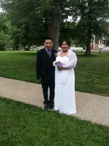 Two people standing on a sidewalk in a park, one in a black suit and one in a white dress holding flowers.