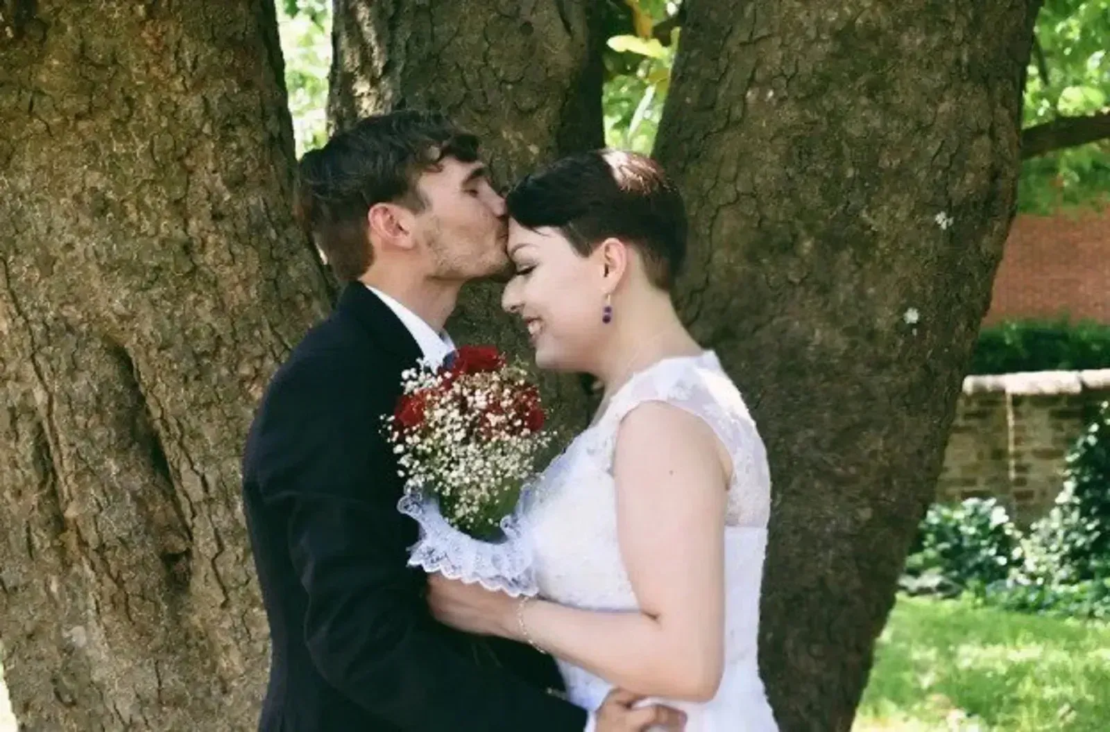 Wedding couple embracing under a tree, groom kissing bride’s forehead, holding a bouquet of red and white flowers