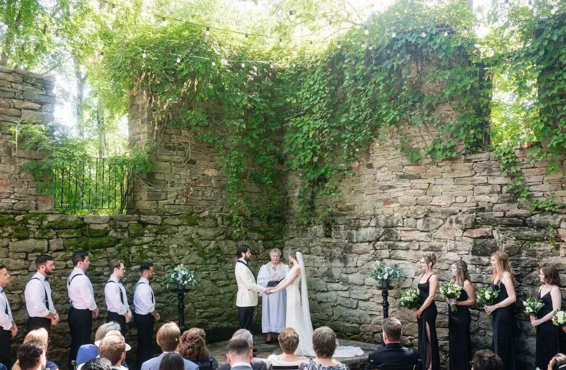 Outdoor wedding ceremony before a stone wall draped in greenery, with wedding party and guests seated.