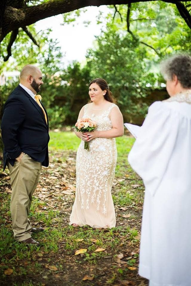 Bride and groom stand outdoors during a wedding ceremony, with an officiant nearby.