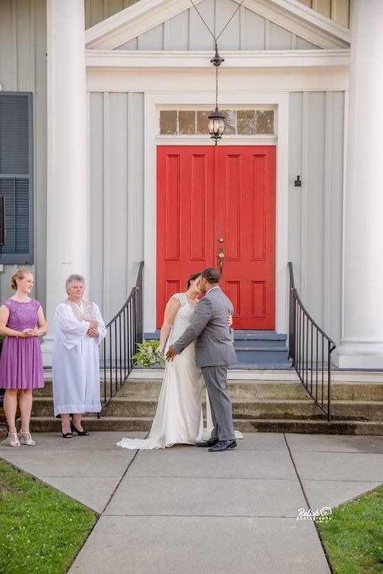 Wedding couple kissing in front of bright red church doors, with guests standing nearby