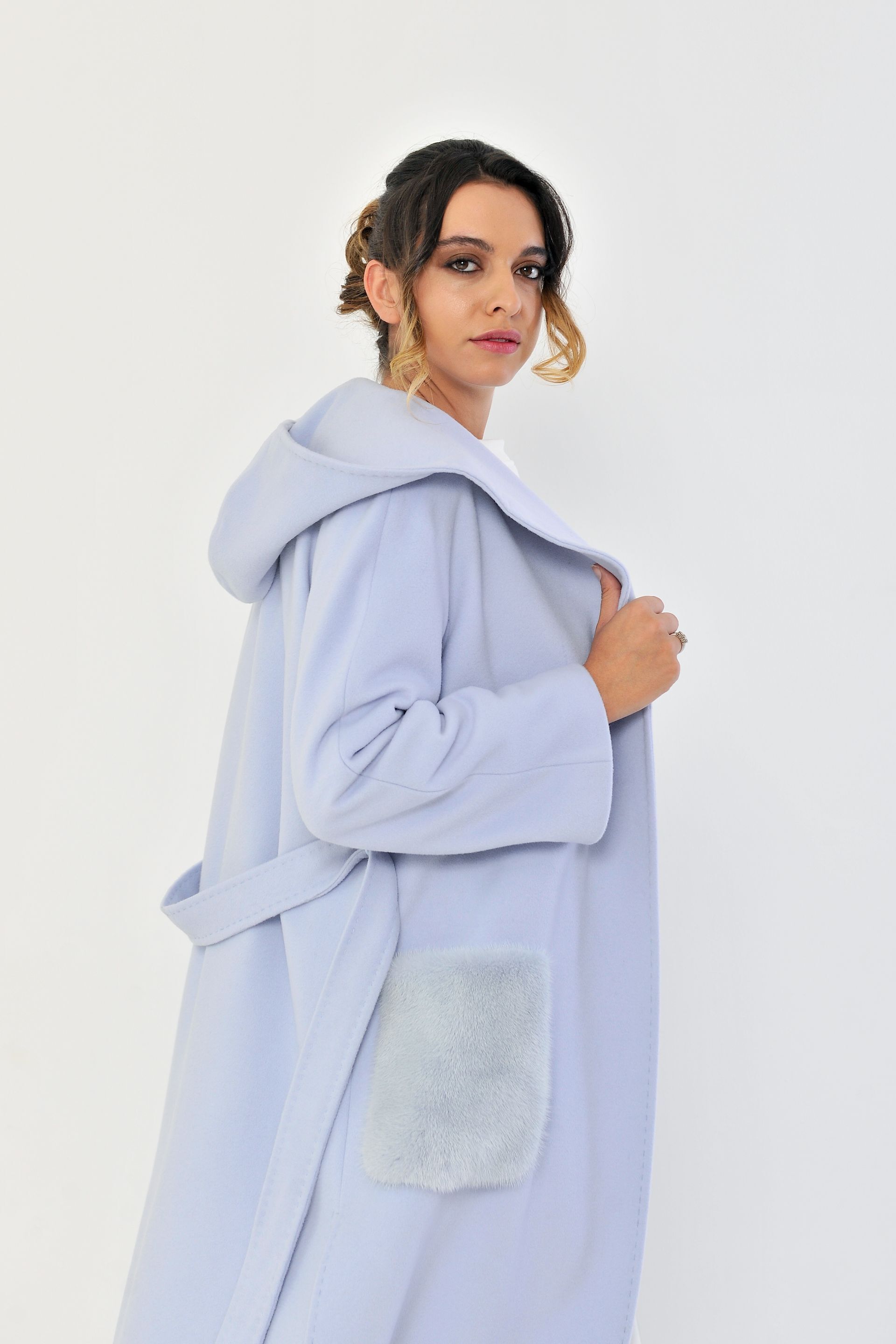 Donna con cappotto azzurro e pantaloni bianchi, in piedi con una mano in tasca contro un muro bianco.