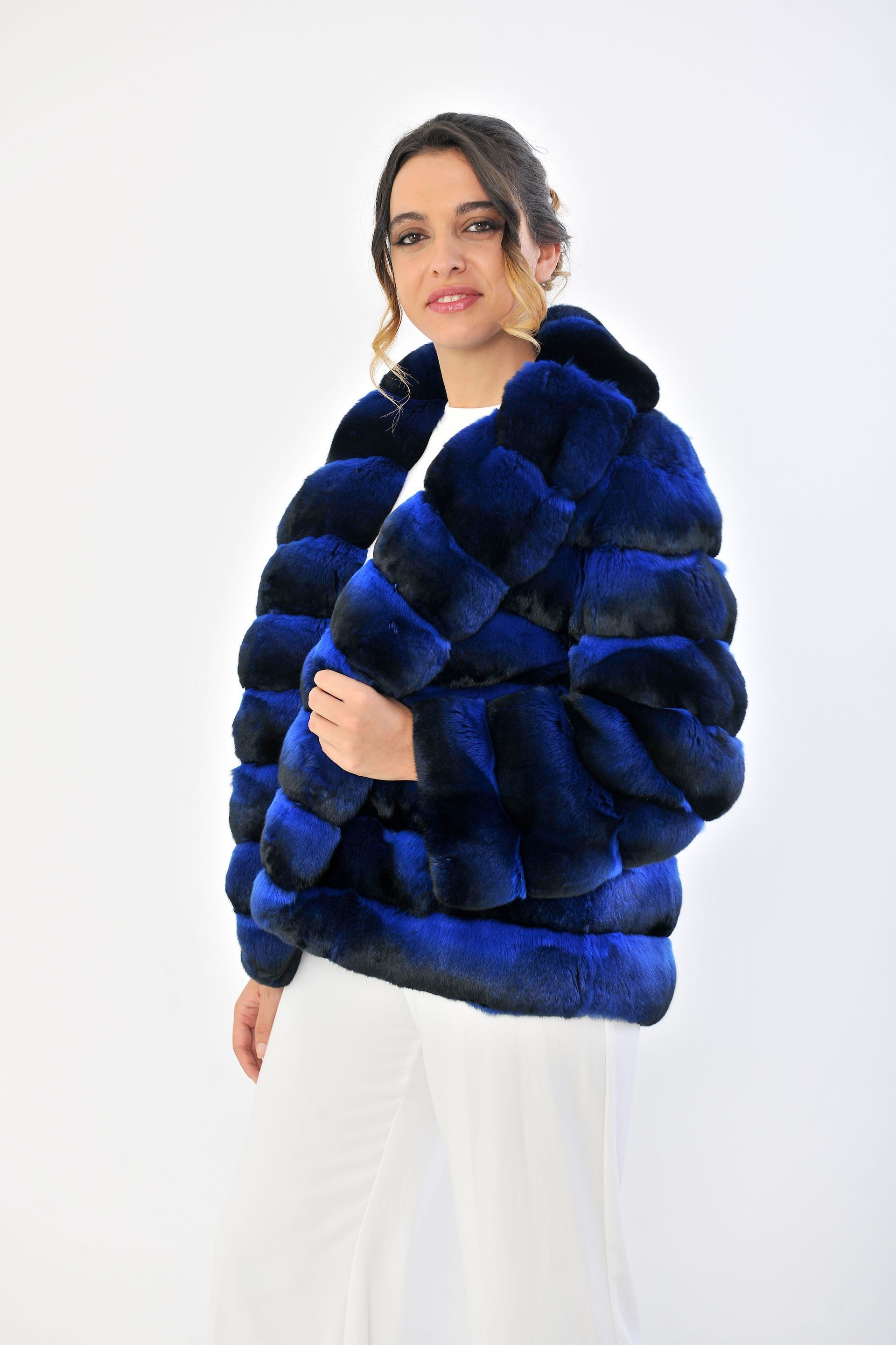 Una donna con una giacca di pelliccia blu e pantaloni bianchi è in piedi su uno sfondo bianco.