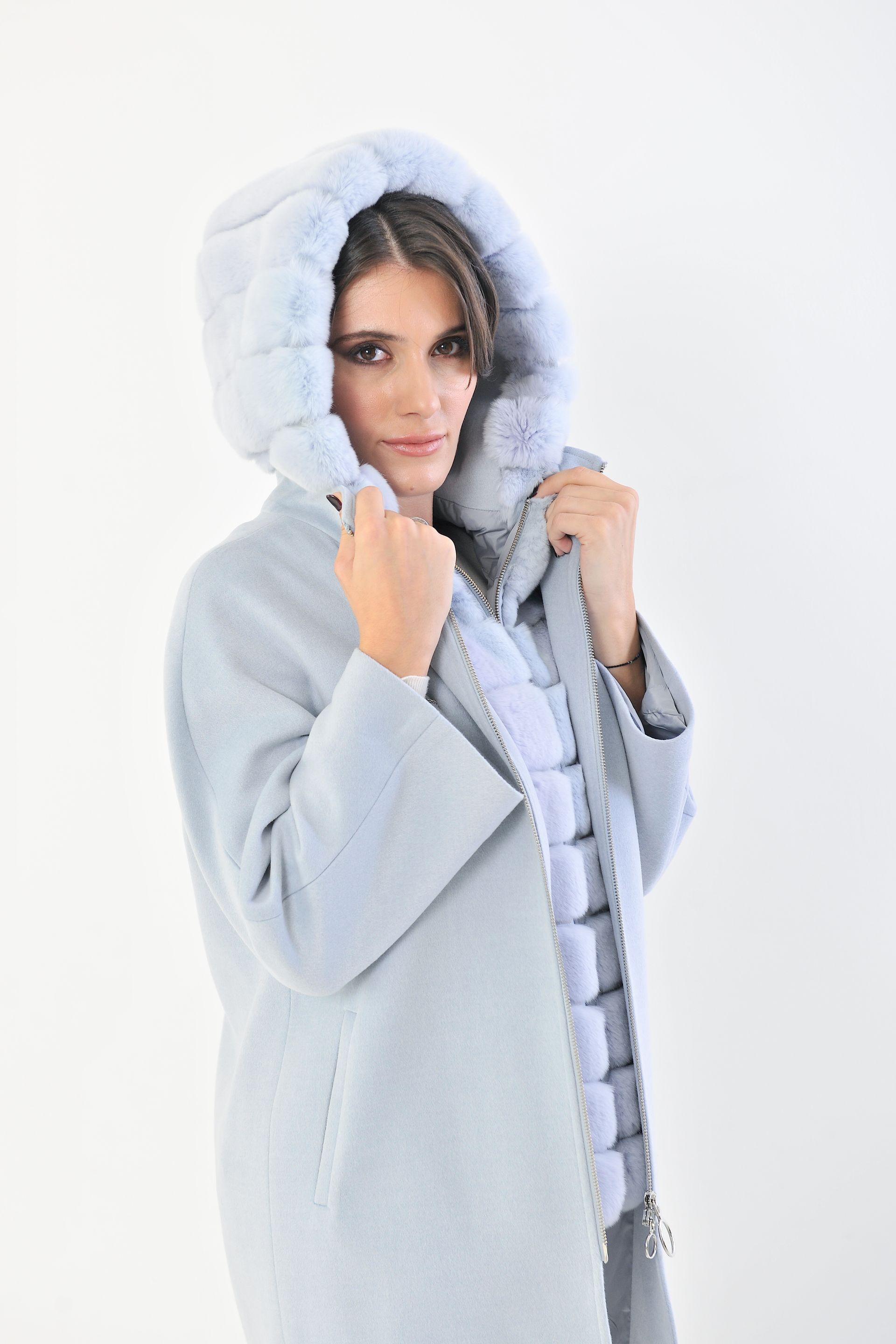 Donna che indossa un cappotto azzurro con rifiniture in pelliccia, pantaloni bianchi, mano in tasca, su sfondo bianco.