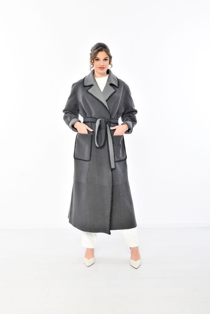 Donna con lungo cappotto grigio con rifiniture nere, in piedi di lato, pantaloni bianchi, tacchi.