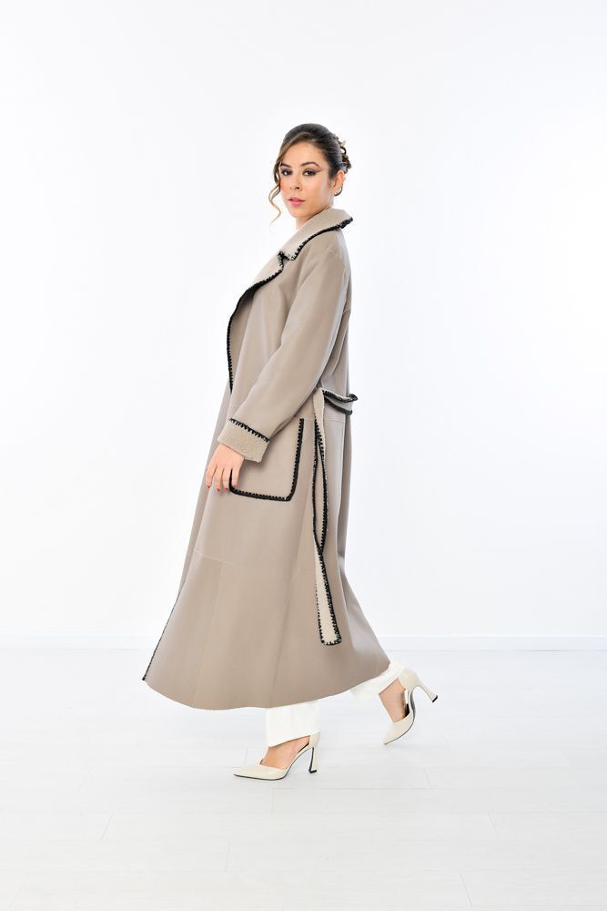 Donna in piedi con cappotto beige con rifiniture e cintura nere e maglione bianco.