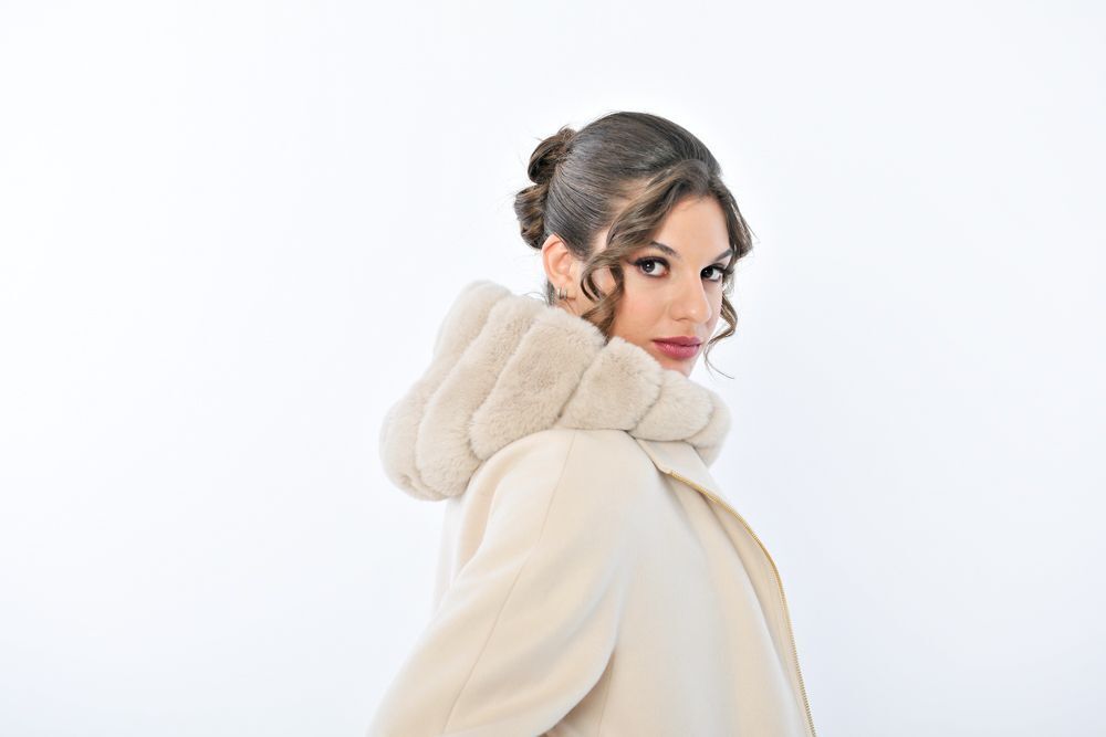 Donna con cappotto color crema e cappuccio bordato di pelliccia, che guarda oltre le spalle, su sfondo bianco.