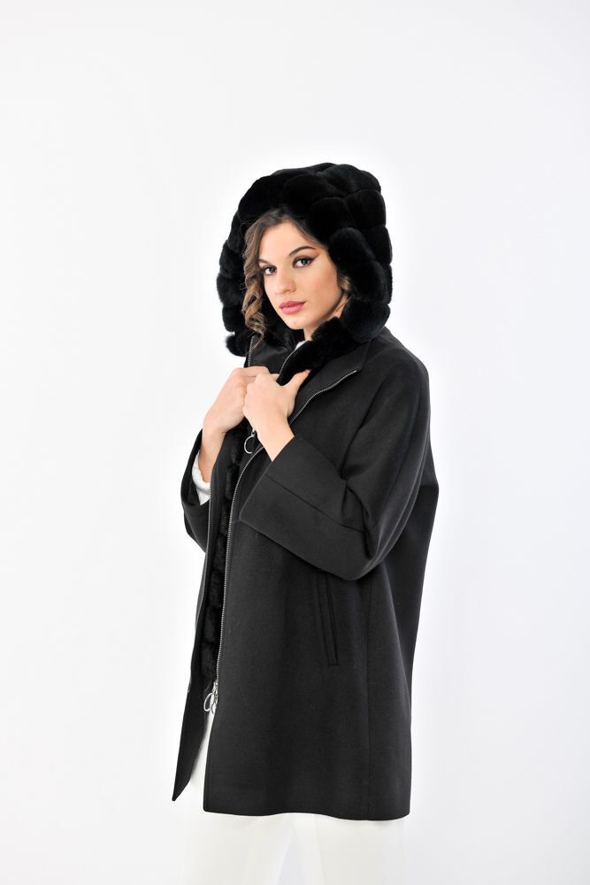 Donna con cappotto nero, cappuccio di pelliccia e pantaloni bianchi, in piedi su uno sfondo bianco.