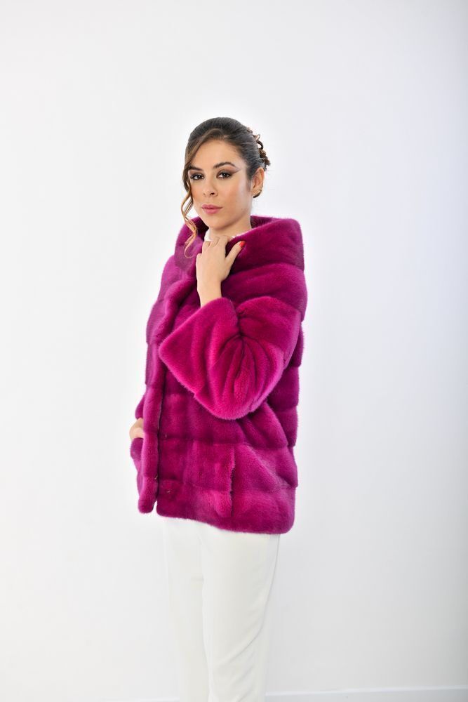 Donna con una giacca di pelliccia rosa acceso con tasche, pantaloni bianchi, in posa su uno sfondo bianco.