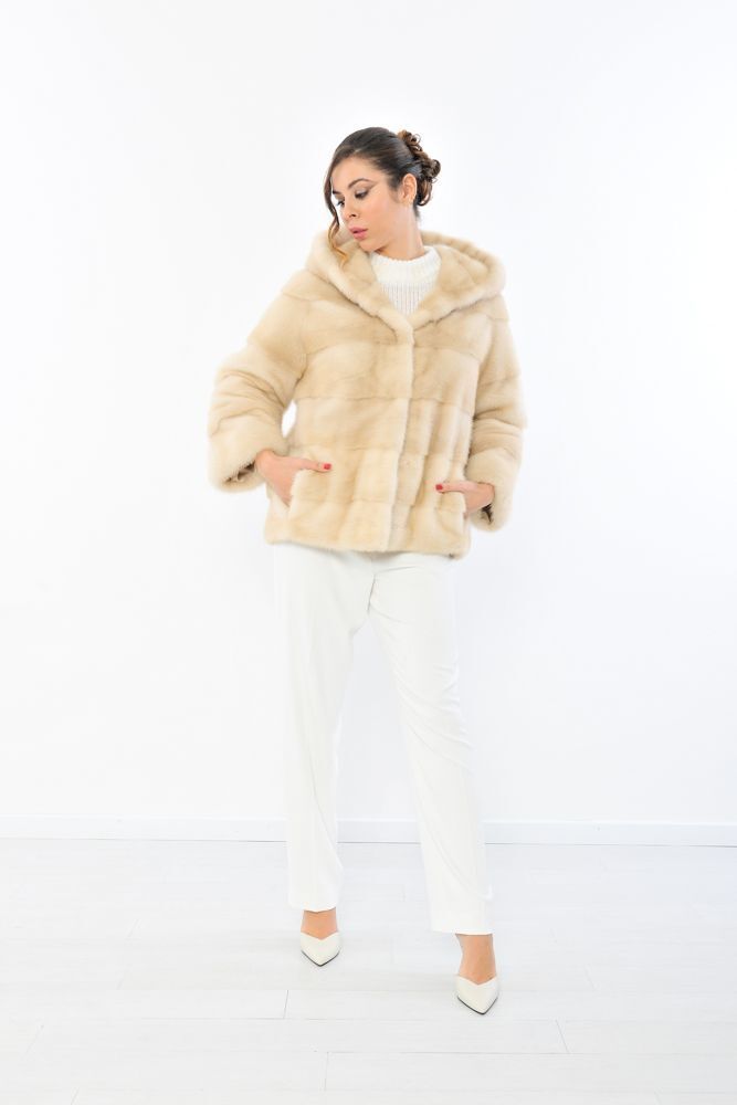 Donna con pelliccia color crema con cappuccio, maglione bianco e pantaloni, su sfondo bianco.