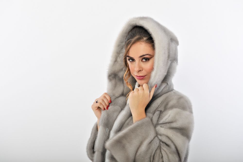 Donna che indossa un cappotto di pelliccia grigio con cappuccio, top bianco e pantaloni, in piedi su uno sfondo bianco.