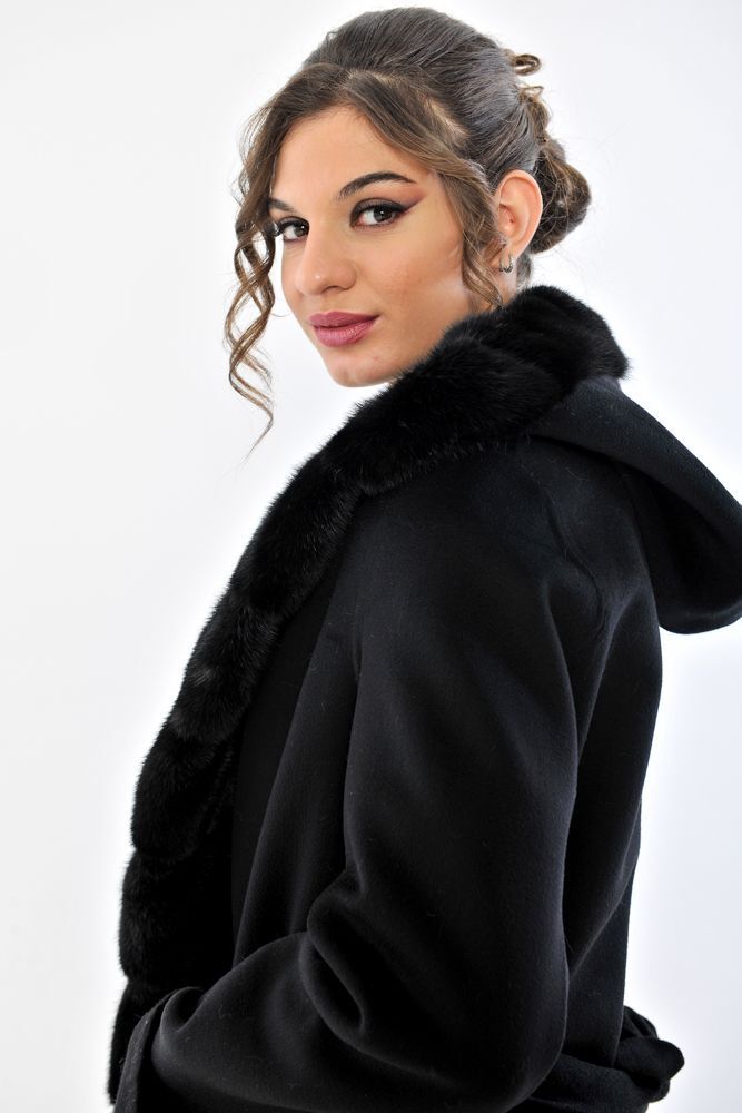 Donna con cappotto nero e cappuccio foderato di pelliccia, mani in tasca, su sfondo bianco.