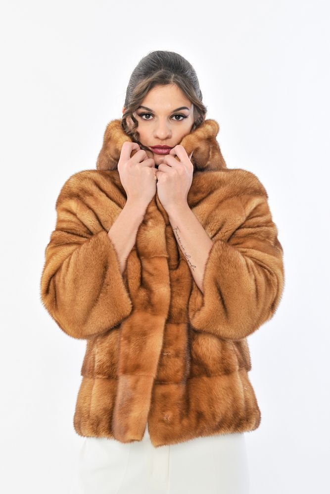 Donna con pelliccia color cuoio, cappuccio e pantaloni bianchi, mani in tasca.