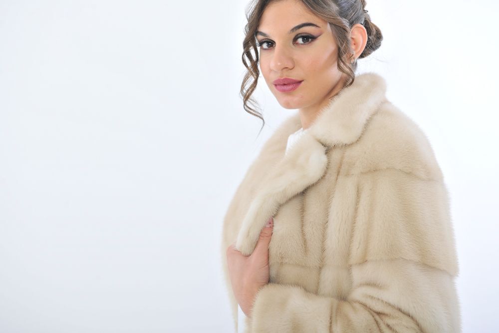 Donna con pelliccia color crema, maglione bianco e pantaloni, in piedi su uno sfondo bianco.