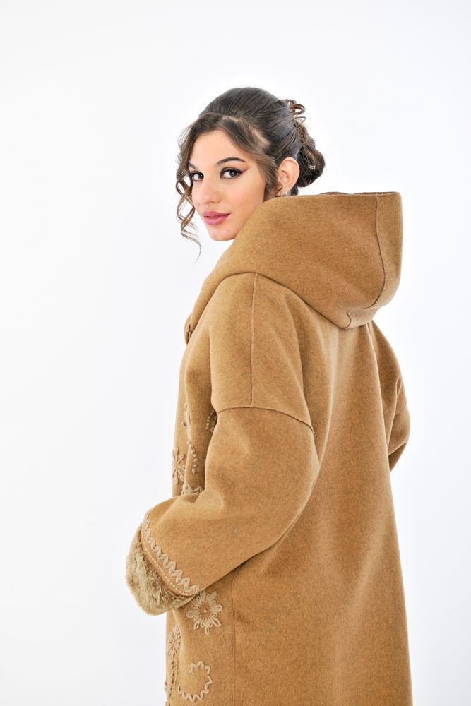 Donna con cappotto marrone chiaro con cappuccio, pantaloni bianchi e tacchi, in piedi in uno studio.