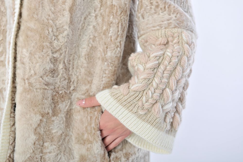 Donna in piedi con le mani in tasca e un cappotto marrone chiaro con cappuccio e rifiniture bianche.