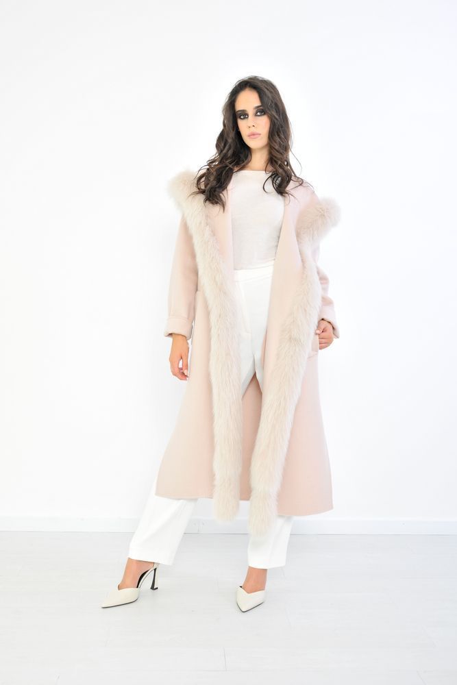 Donna con cappotto rosa foderato di pelliccia e pantaloni bianchi, in posa in un interno con sfondo bianco.