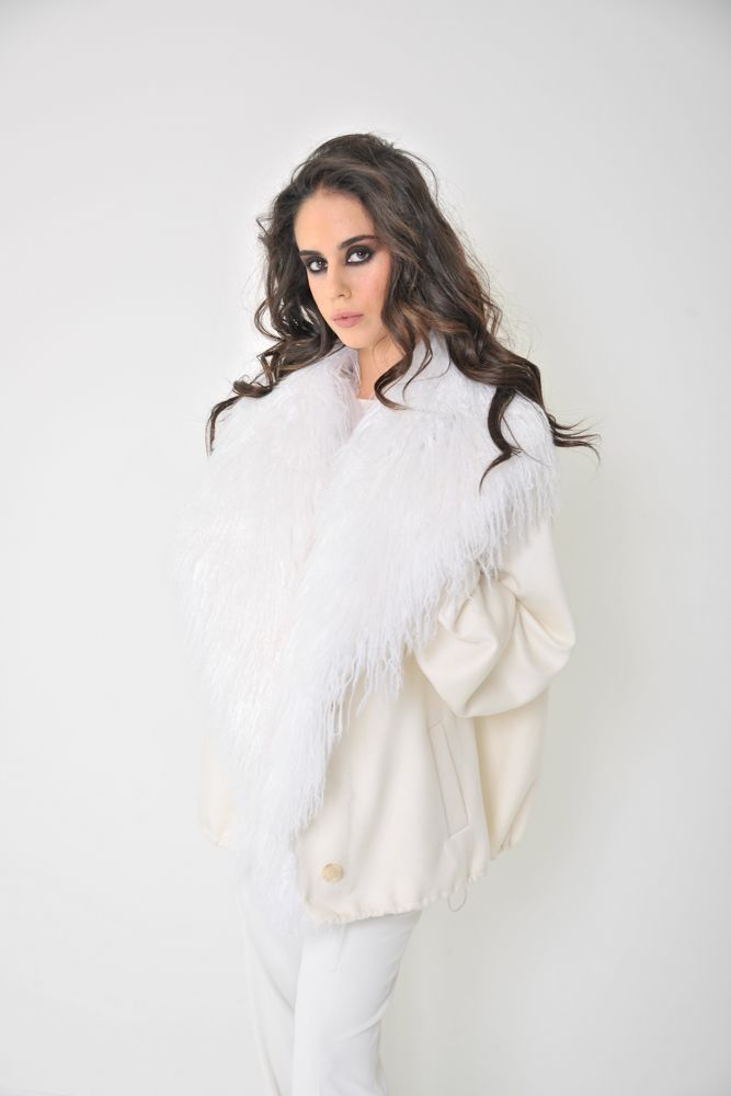 Donna in abito bianco con colletto di piume, su sfondo bianco. Ha i capelli scuri e un'espressione seria.