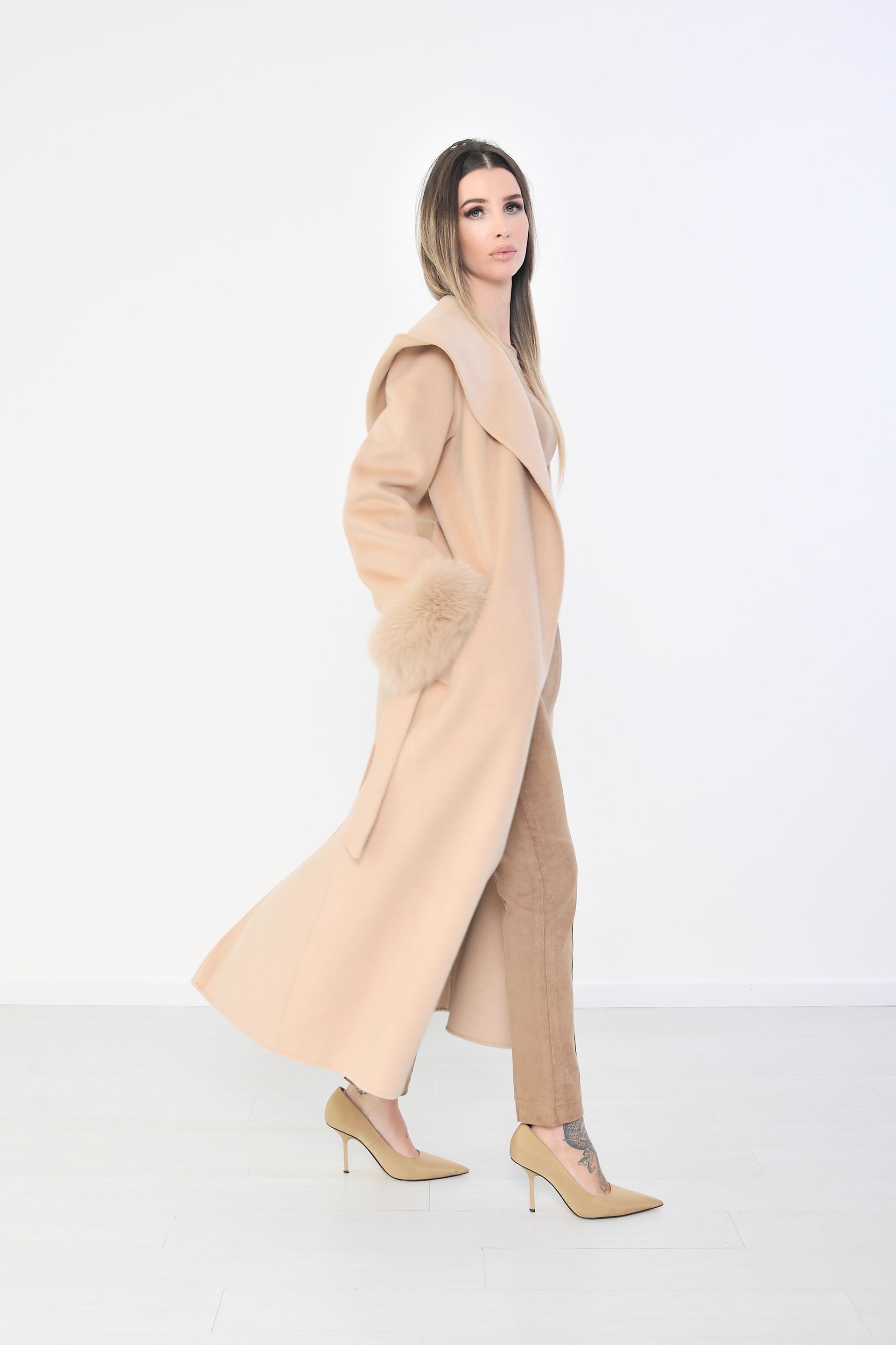 Donna con cappotto marrone chiaro con polsini di pelliccia, cappuccio alzato, in posa davanti a uno sfondo bianco.