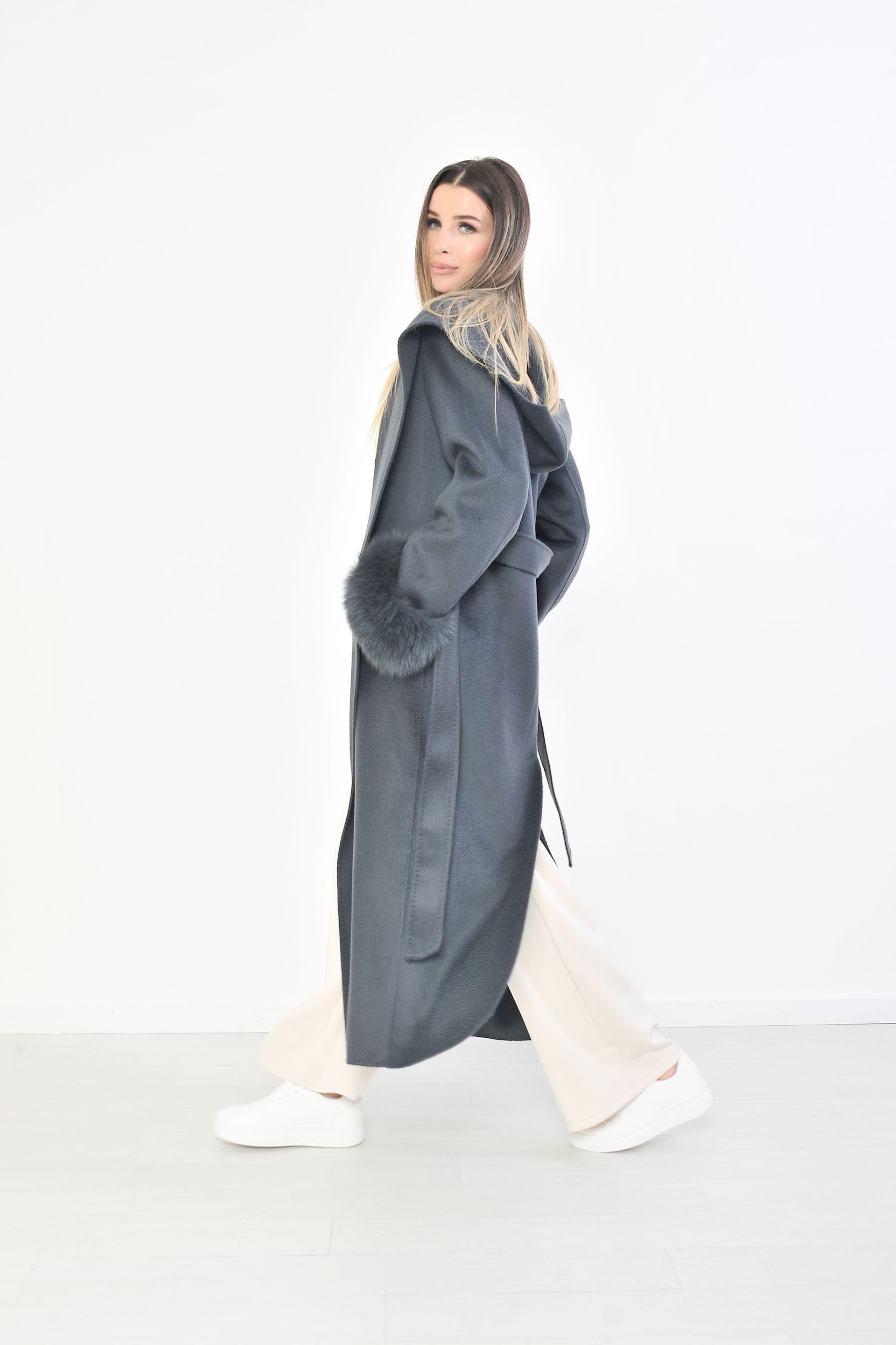 Una donna con un cappotto grigio, polsini e cappuccio di pelliccia, è in piedi davanti a uno sfondo bianco.