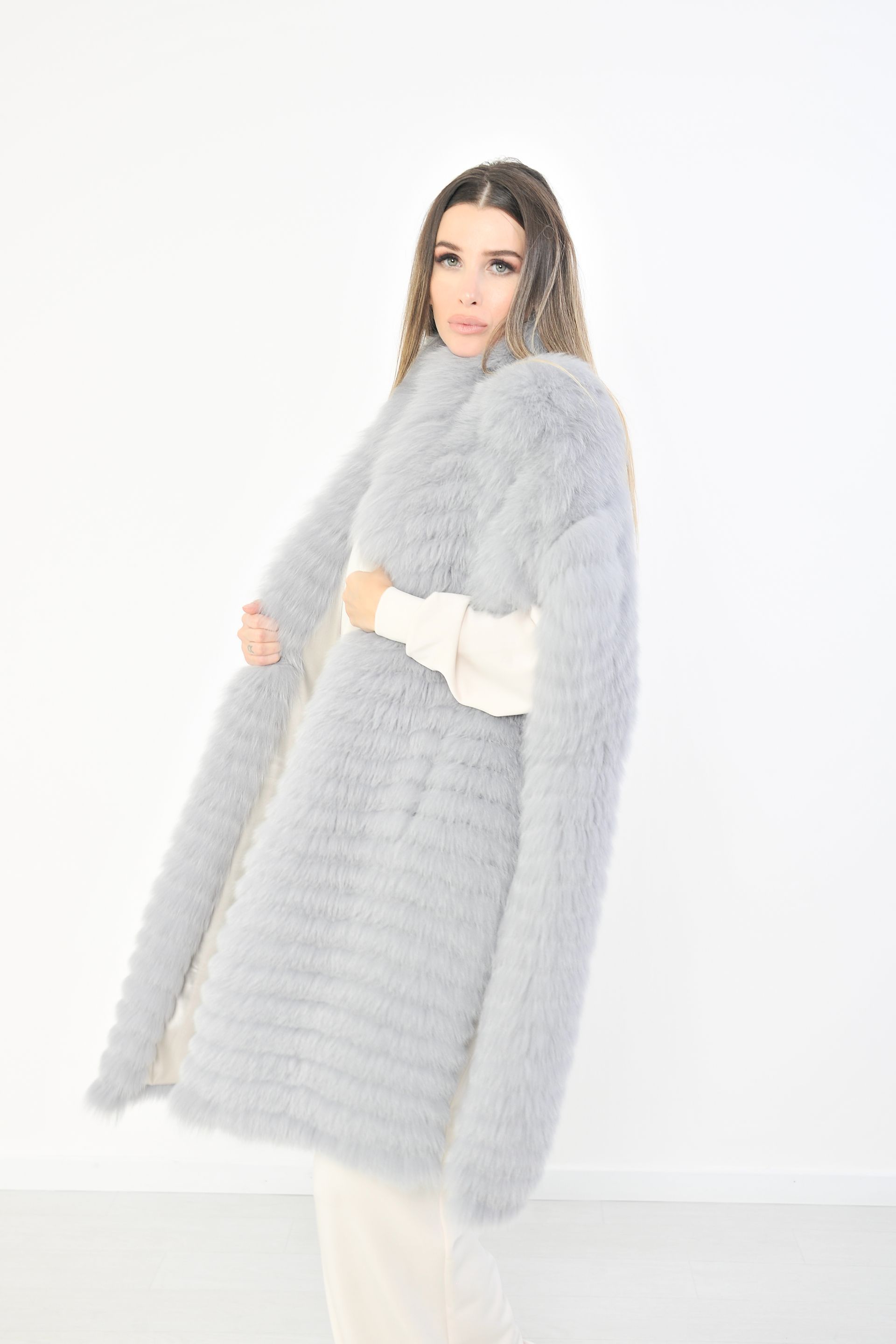 Donna con pelliccia azzurra e maglione bianco, sorride su uno sfondo bianco.