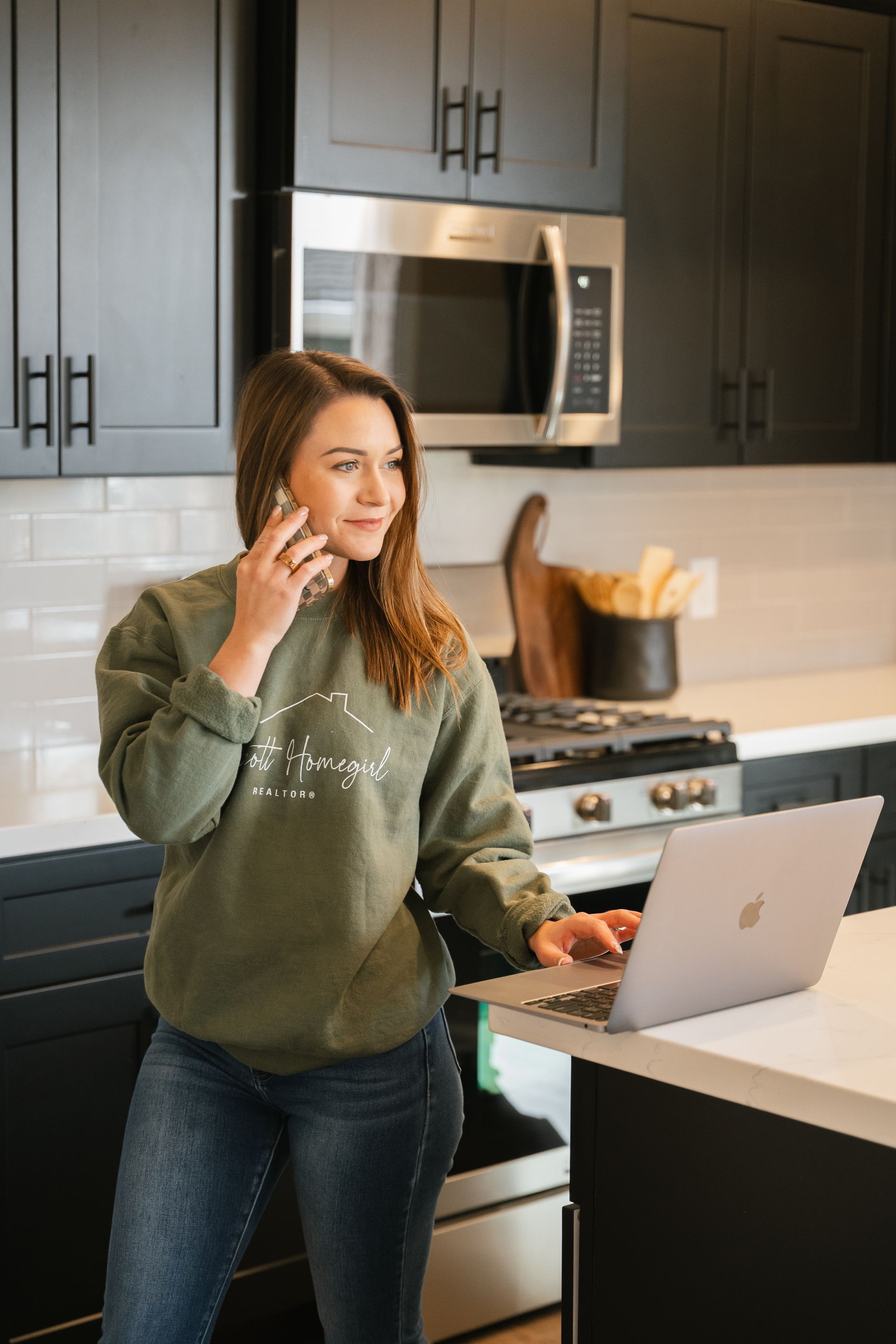 Bree is standing in a kitchen talking on a cell phone while using a laptop computer.