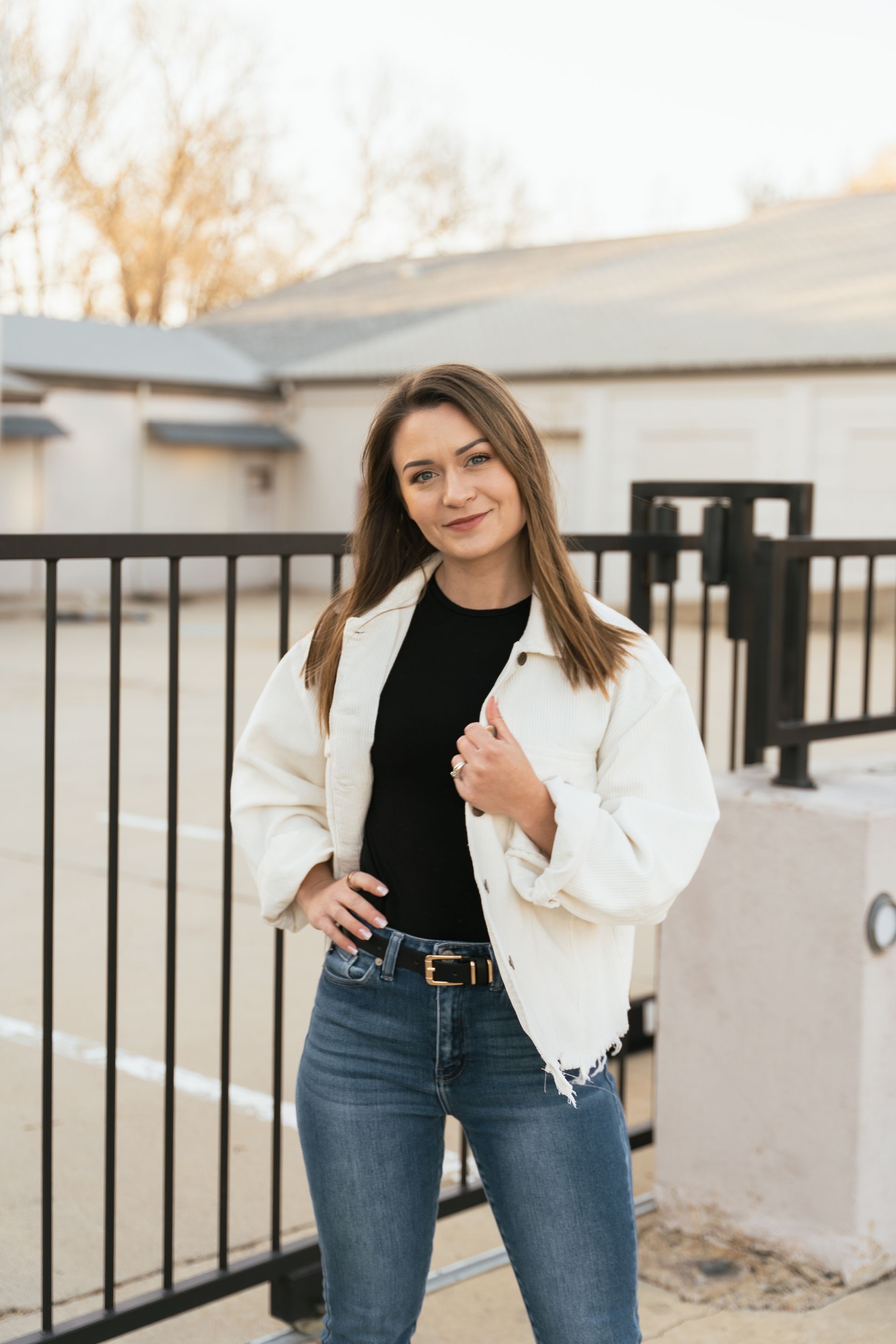 Bree wearing a white jacket and jeans is standing in front of a fence.