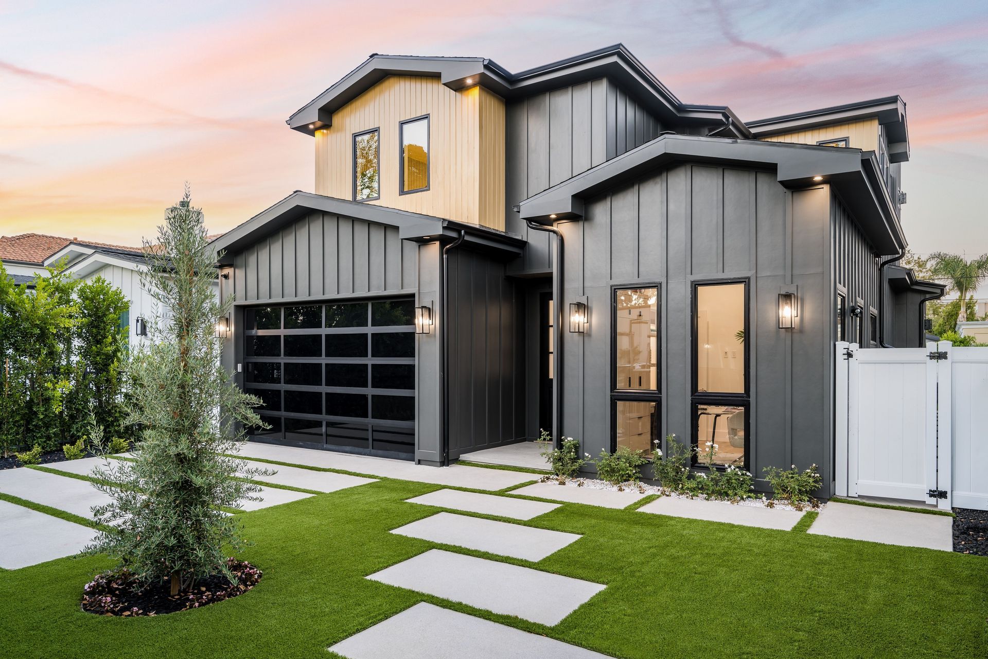 Modern two-story house with dark gray siding, glass garage door, and a green lawn with stone walkway.