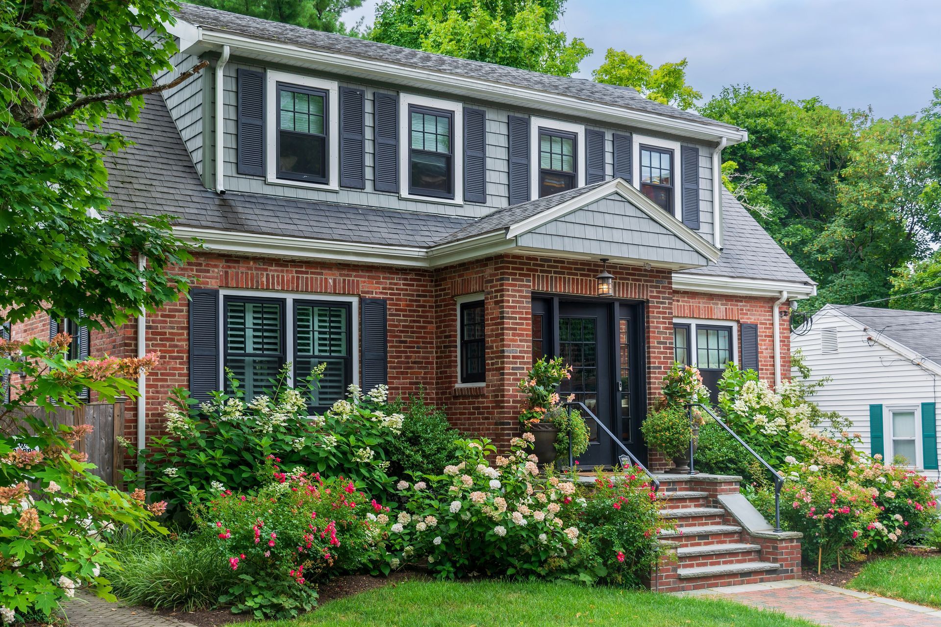 Red brick house with dark shutters, front porch, and lush landscaping.