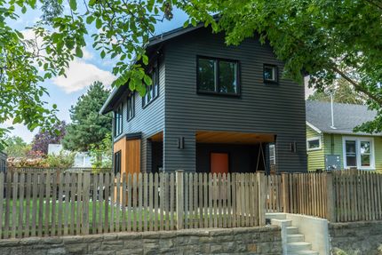 Modern black house with wooden accents and orange door, behind a wooden fence.