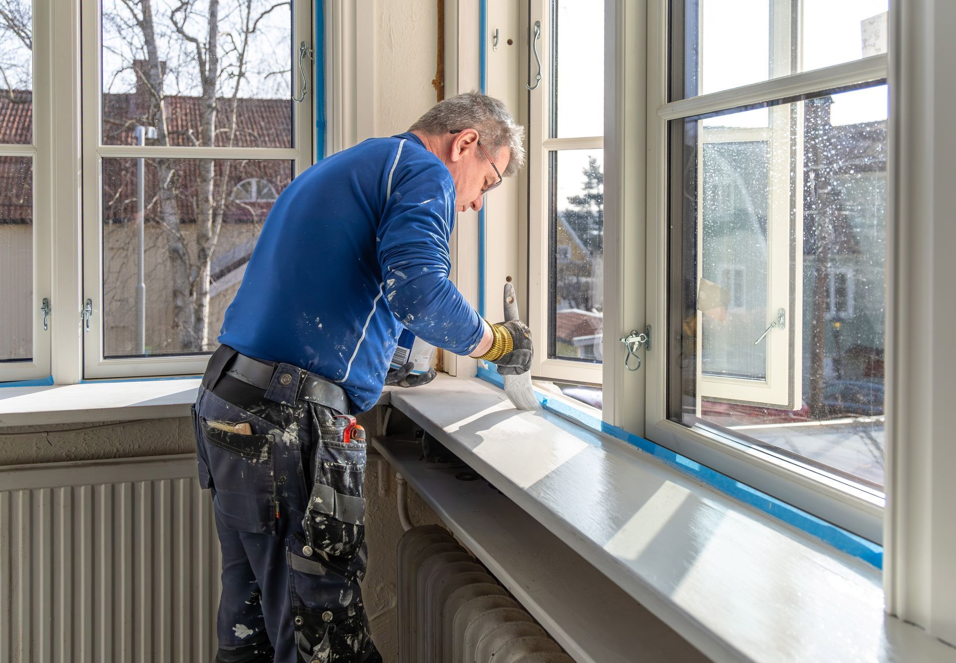 A person in blue painting a white windowsill near a window. The room has a radiator and tape on the window frame.