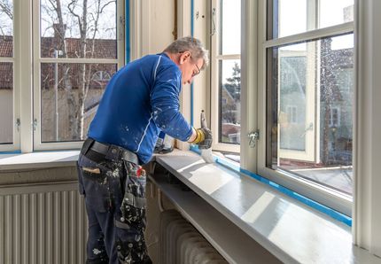 A person painting a white windowsill inside. Blue painter's tape protects window, sunny room.