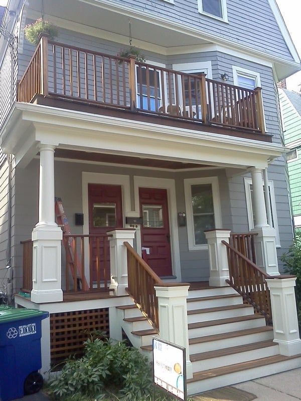 Two-story house with a front porch and balcony. Light gray exterior, wooden railing, and red doors.