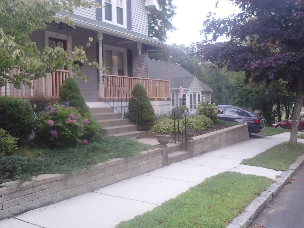 A two-story gray house with a porch and steps, featuring landscaping and a sidewalk.