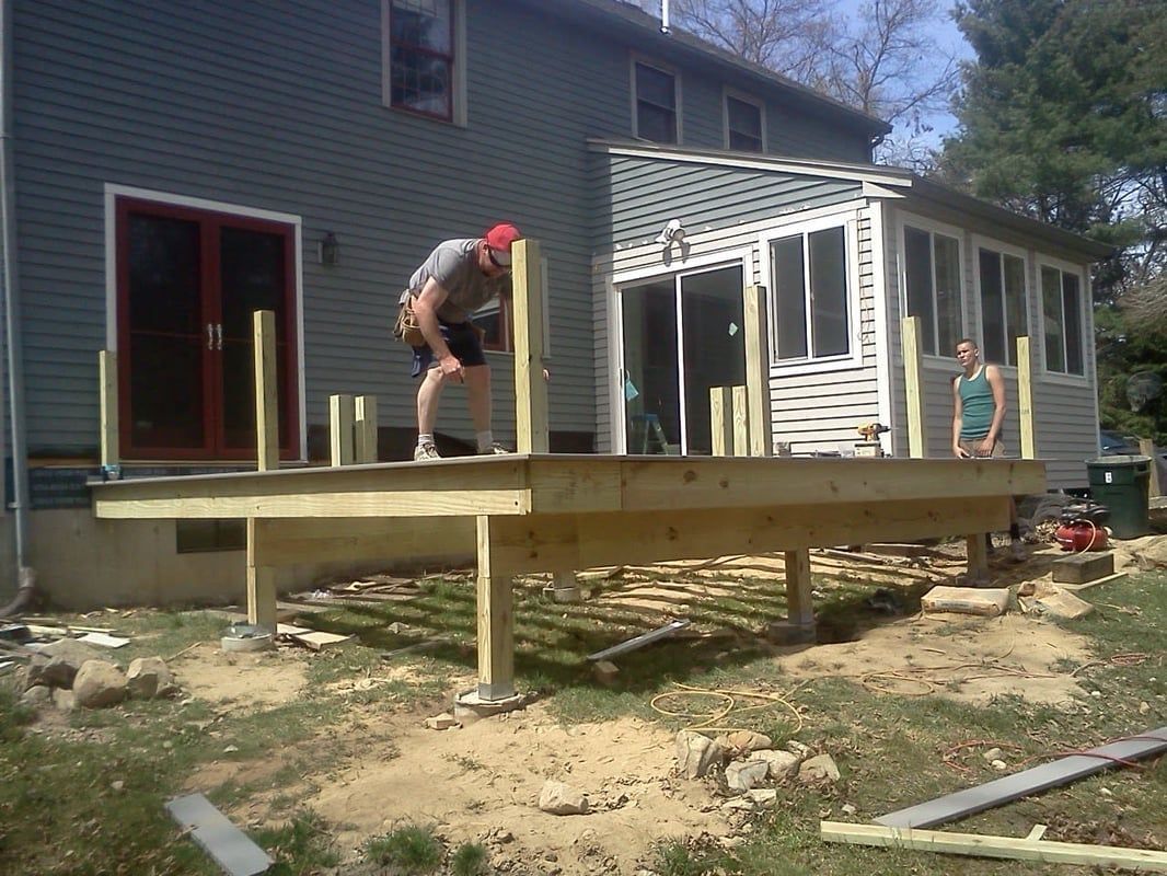 Construction of a wooden deck; two people working on it next to a house with a sunroom.