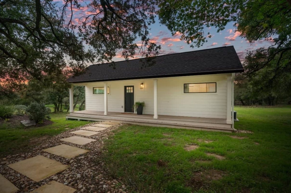 White house with black roof, small porch, stone path, green lawn, trees, and colorful sunset.