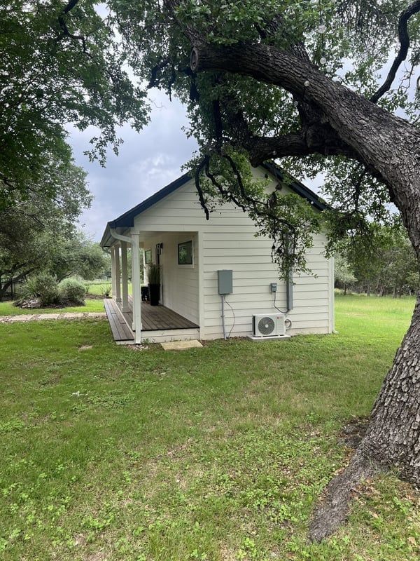 Small white cabin with porch, shaded by a tree, set in a grassy yard.
