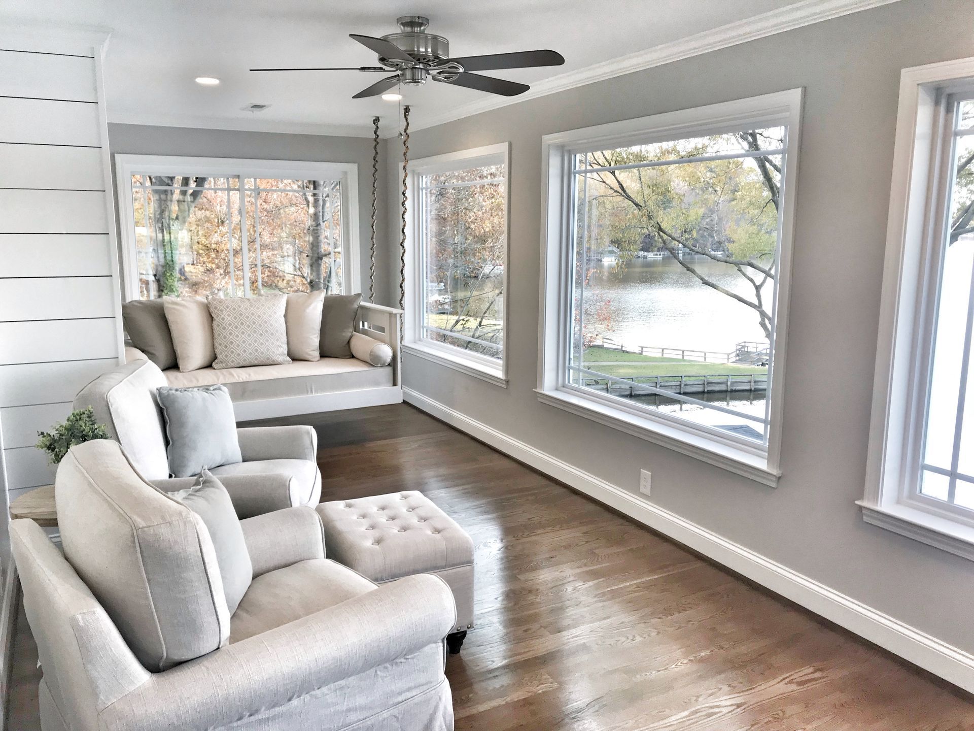 Sunroom with gray walls, wood floor, white furniture, a swing, and large windows overlooking a lake.