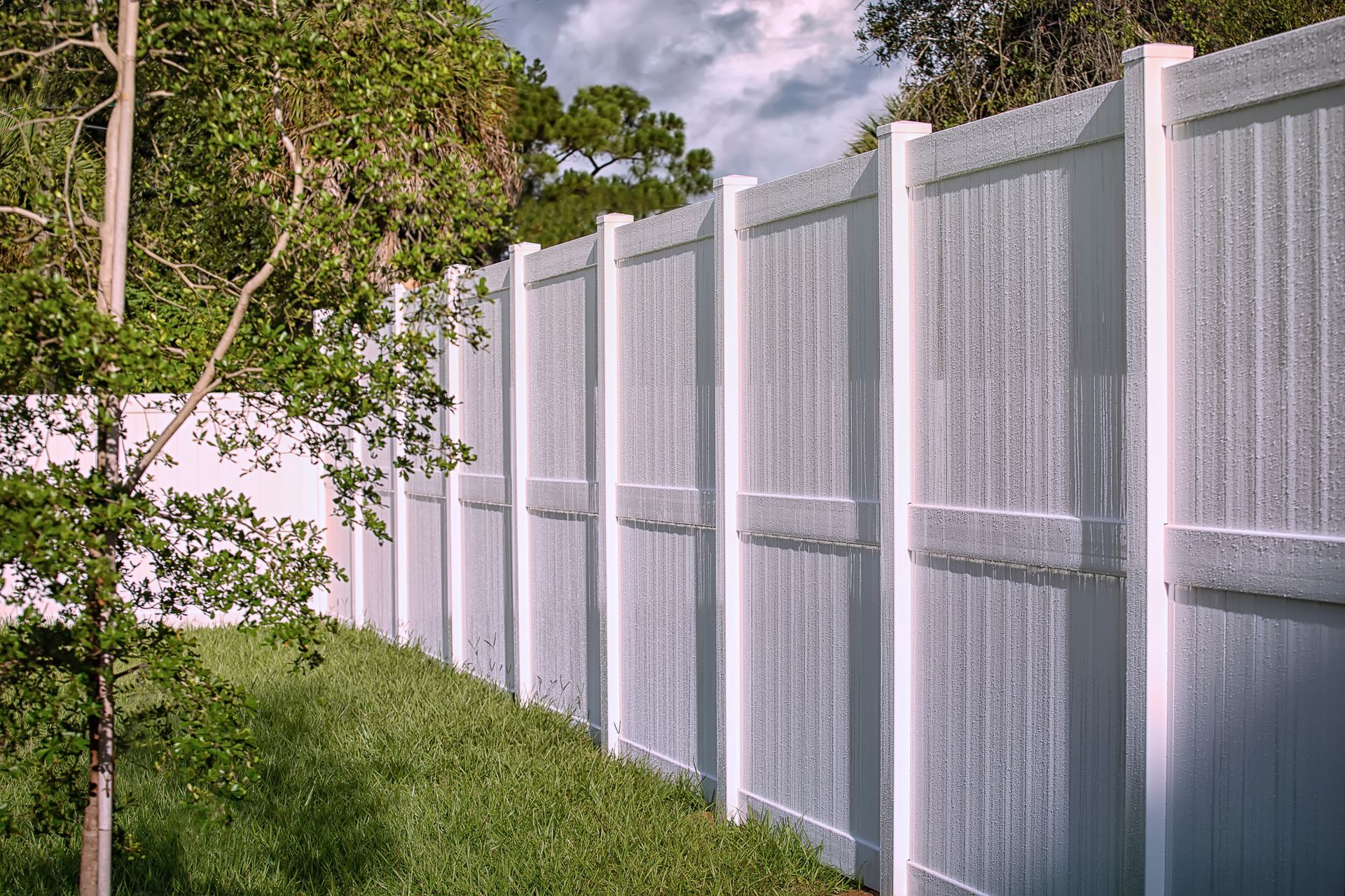 White vinyl privacy fence in a grassy yard, trees in background.