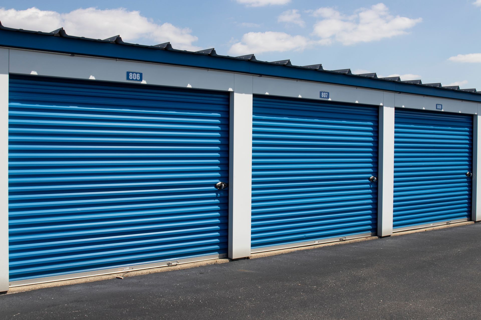 Blue storage unit doors in a row; exterior, asphalt lot, sunny day.