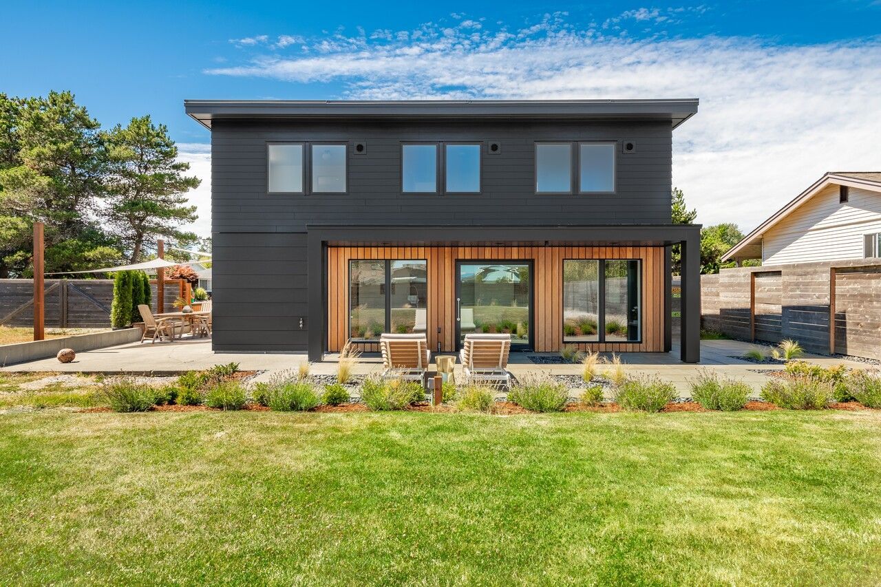 Modern black house with wood accents, large windows, and outdoor seating on a sunny day.