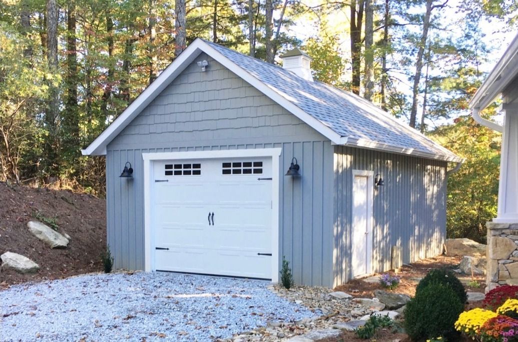 Blue-gray garage with white door, set in a wooded area, on a gravel driveway.