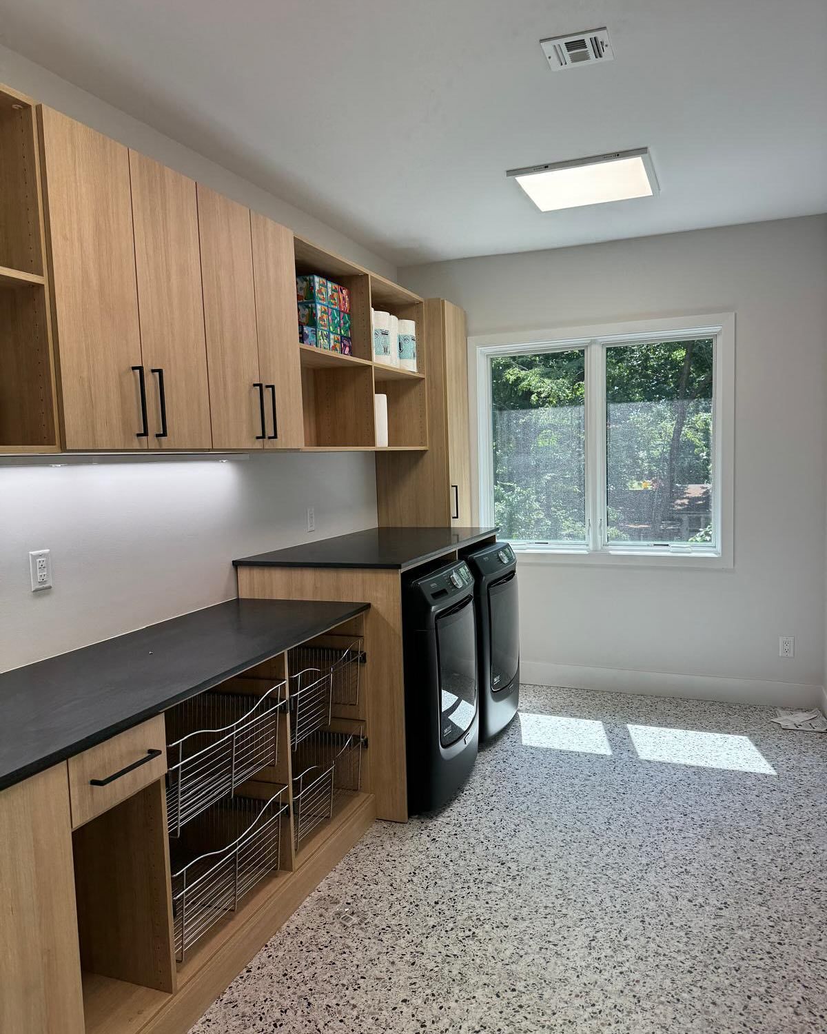 Laundry room with light wood cabinets, black countertops and appliances, speckled flooring, and window.