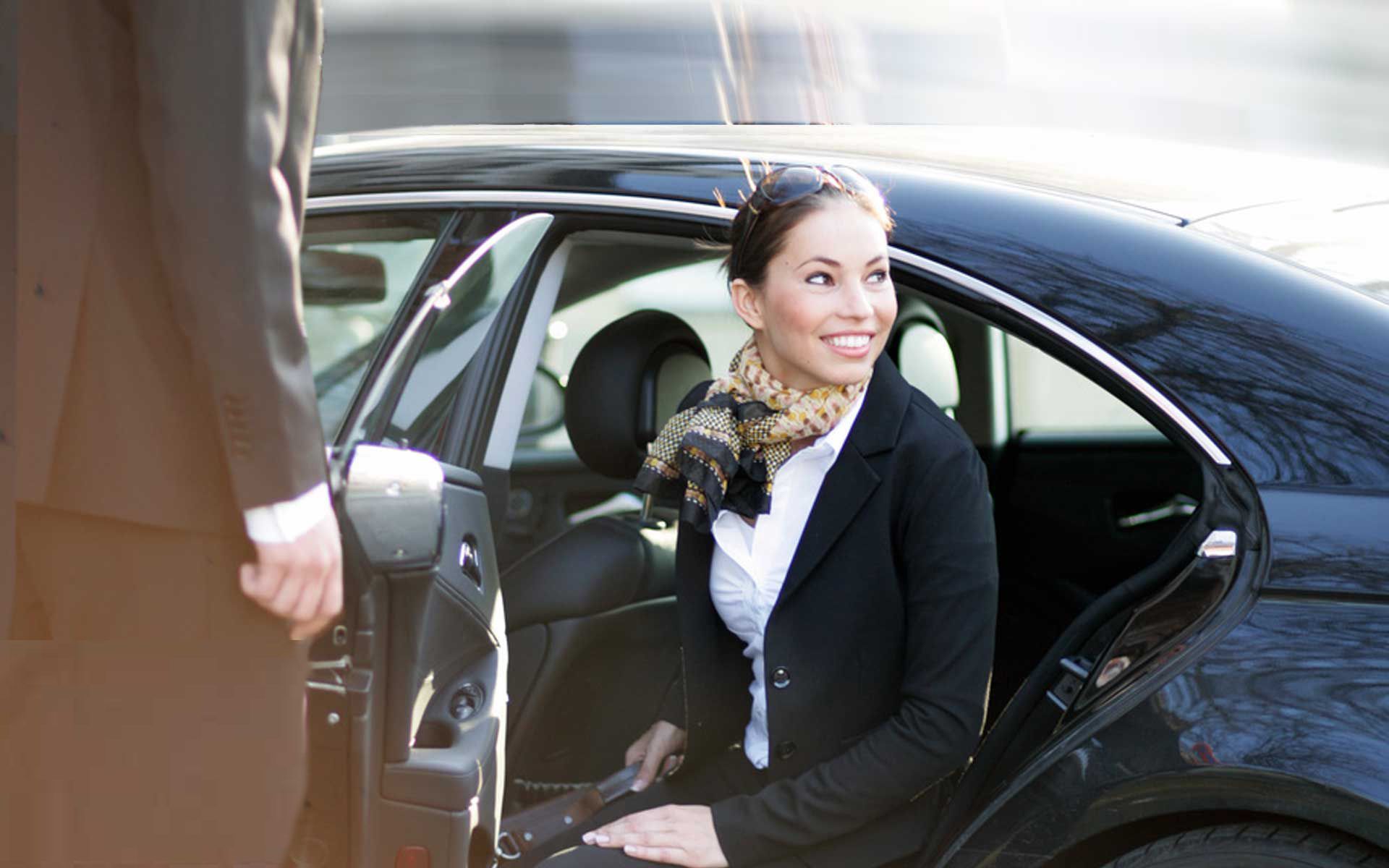 Woman exiting a black car, assisted by a person in a suit. Smiling, outdoors.