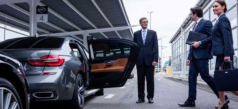 A chauffeur opens a car door for two businesspeople walking towards it at an airport.