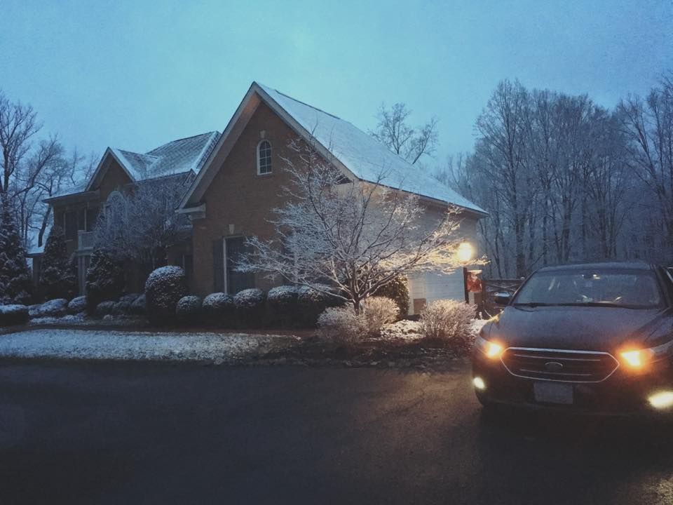 A car with headlights on drives toward a snow-covered house on a winter evening.