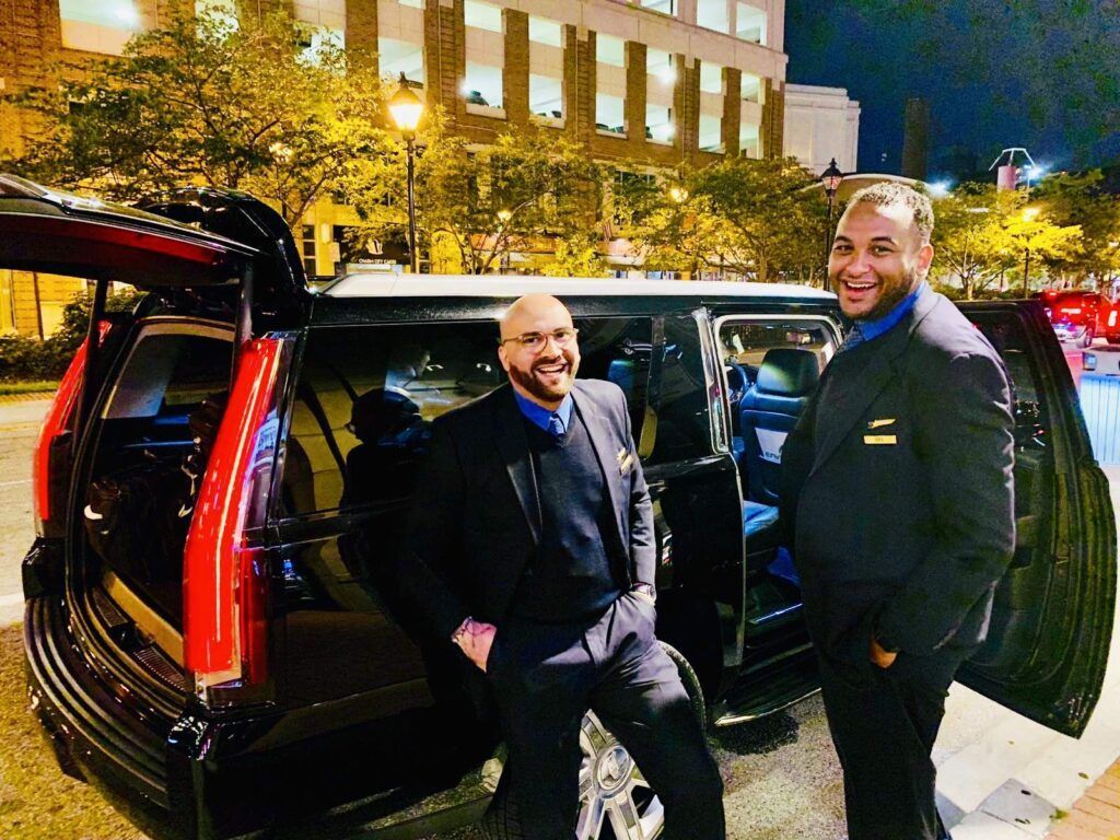 Two men in black suits stand by a black SUV with open doors at night. City background.