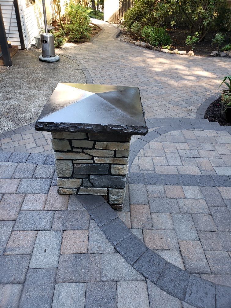 Stone retaining wall with a square pillar and a long ledge, surrounded by greenery and paving stones.