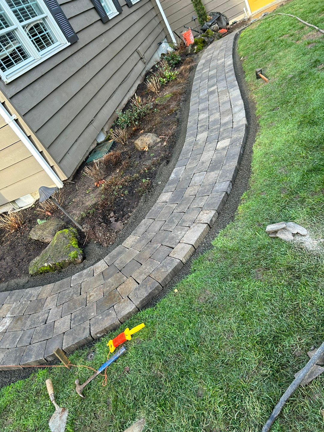 Person laying gray paving stones in a sandy area, wearing gloves and sneakers.