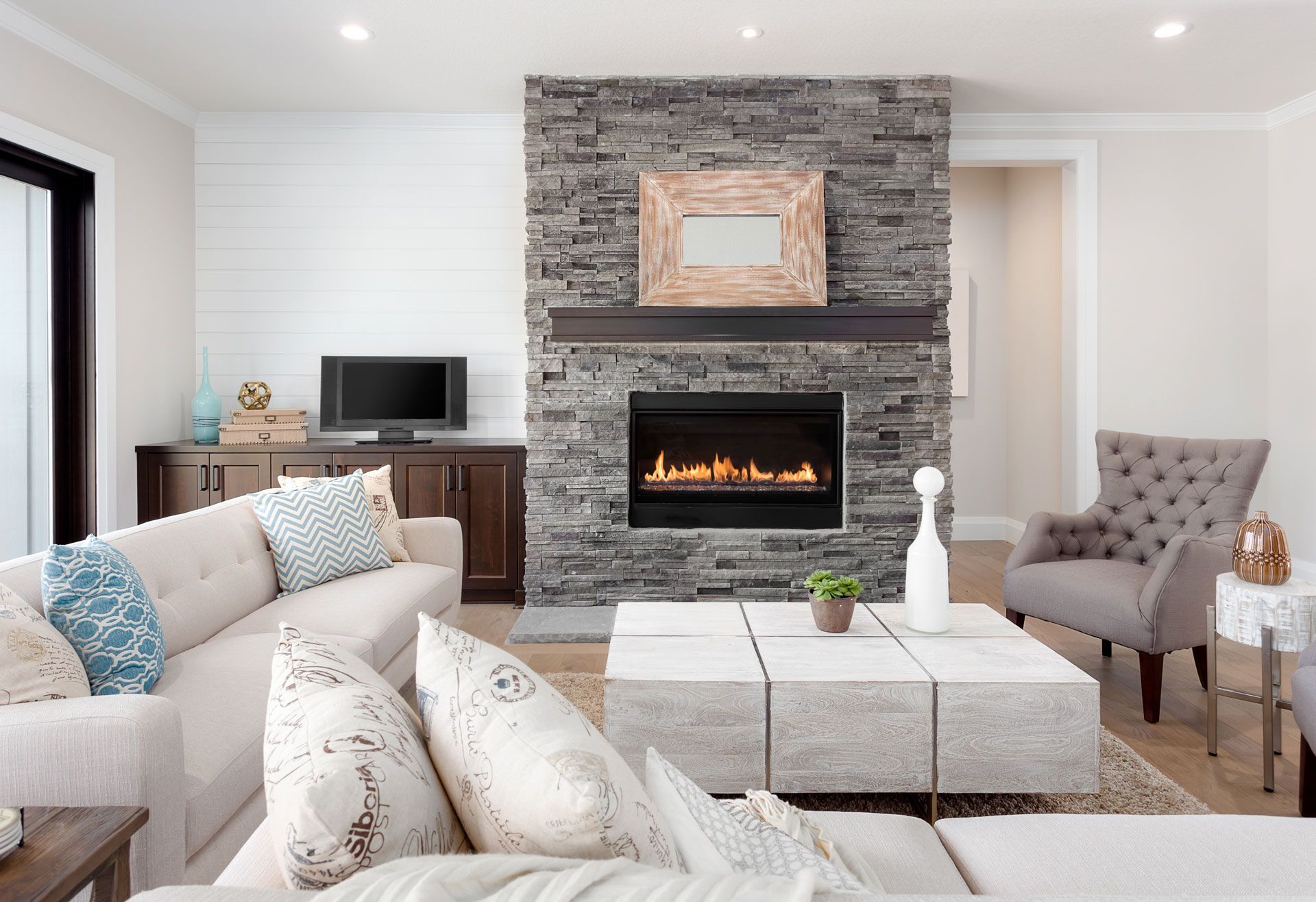 Cozy living room with fireplace and stone wall, light sofa, grey chair, and modern coffee table. Cozy living room with fireplace and stone wall, light sofa, grey chair, and modern coffee table.